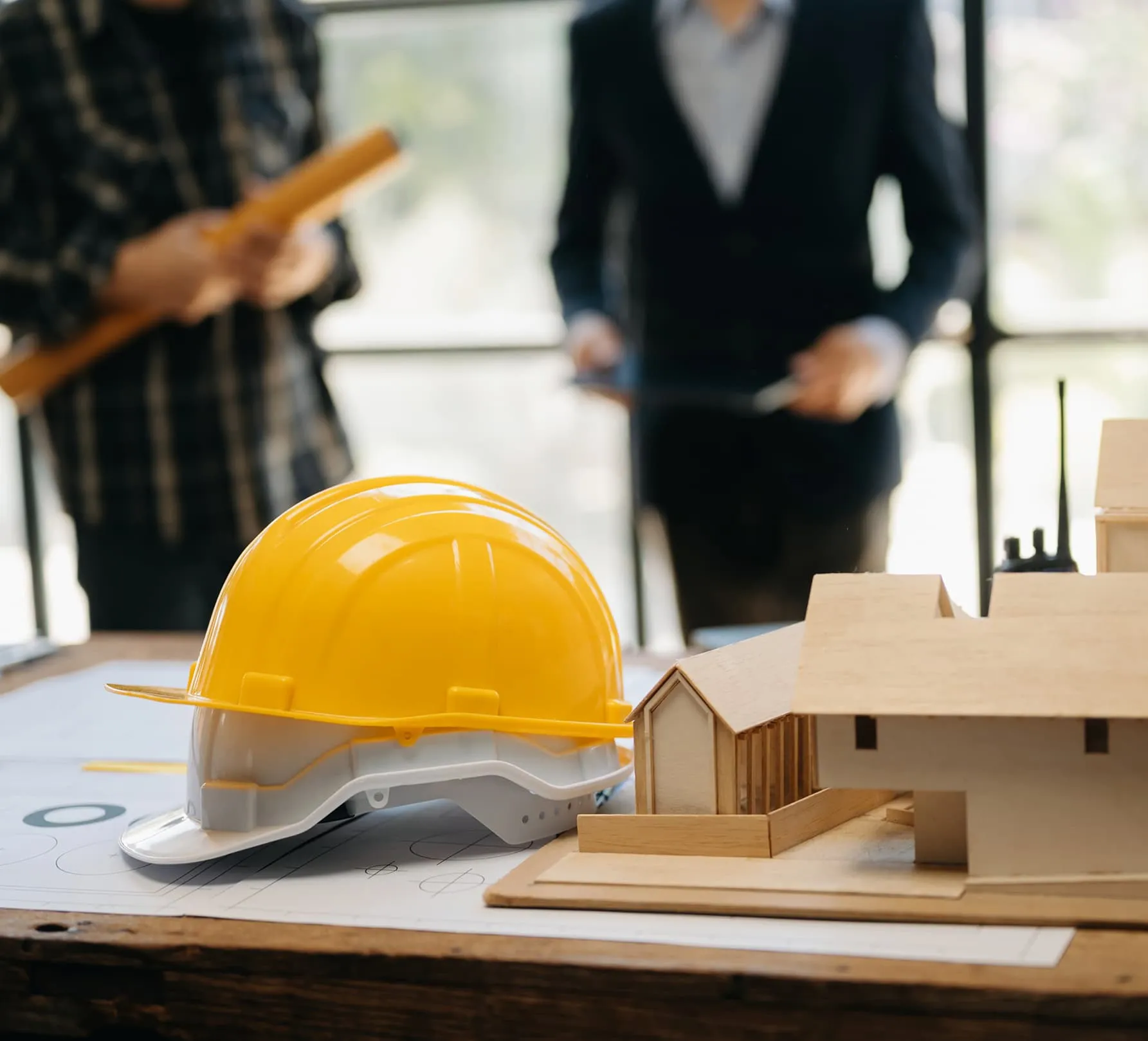 Architectural model and construction helmet on a table, with blurred figures in the background